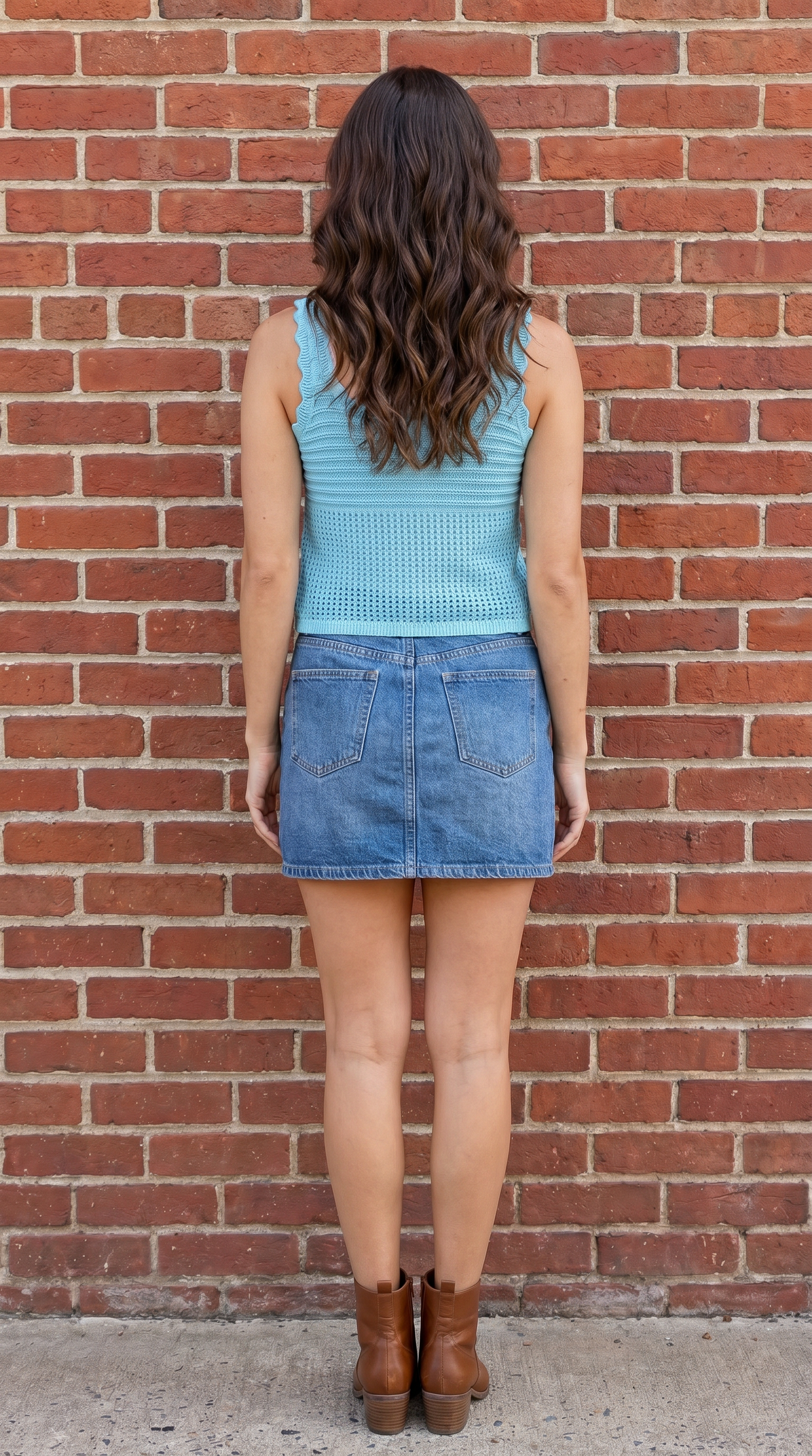 Person wearing a light blue sleeveless top and denim skirt standing against a brick wall.